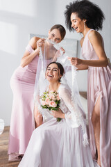 Naklejka premium excited interracial women covering overjoyed bride with veil in bedroom.