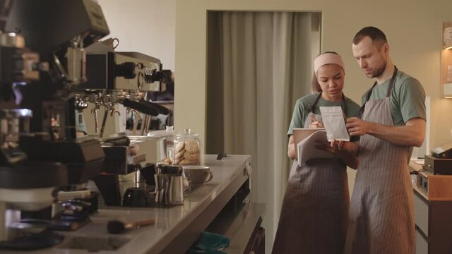 Medium shot of middle-aged Caucasian male barista and his new female mixed-race colleague standing in cafe and discussing coffee pack. Woman making notes in notepad