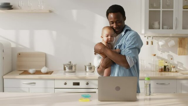 Paternity leave and business. Happy african american father luling newborn baby and working on laptop at kitchen
