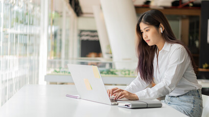 Young Asian students wearing headphones using their laptop in a coffee shop, writing notes on language learning, watching online webinars, audio listening courses, e-learning ideas.