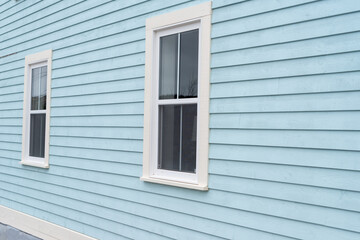 The exterior wall of a baby blue country style house with narrow wood cape cod siding. There are two small double hung windows with white trim. Lace curtains are hanging in the closed glass windows.