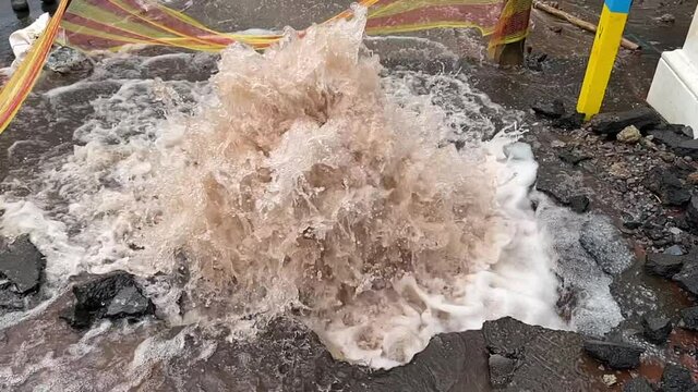 Close up slow-motion of water gushing out of a burst pipe in a damaged city road.