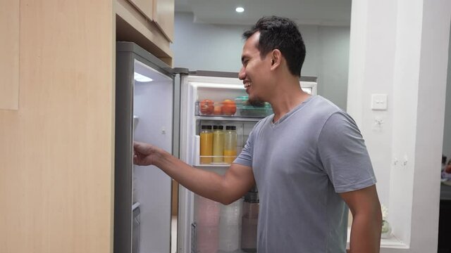 portrait of starving young asian man hold his belly while open the fridge door looking for something to eat
