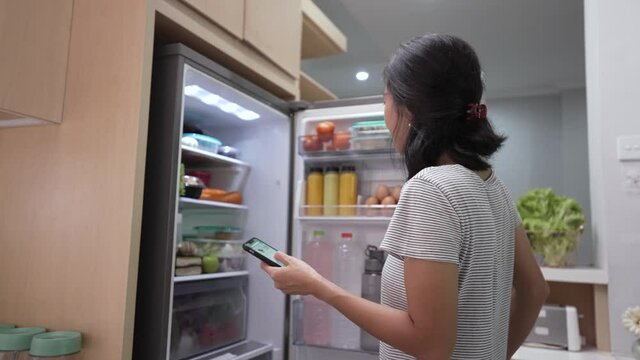 asian young woman using her smartphone to buy groceries while open her fridge at home