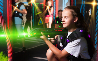 Excited girl aiming laser gun at other players during lasertag game in dark room..