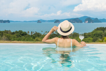 Fototapeta na wymiar Happy woman in white swimsuit drinking wine in luxury swimming pool hotel against beautiful view, young female with hat enjoy in tropical resort. Relaxing, summer travel, holiday and vacation concept
