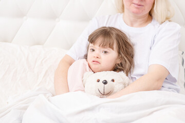 Mom hugging a sleeping girl with down syndrome holding a teddy bear at home in the bedroom on the...