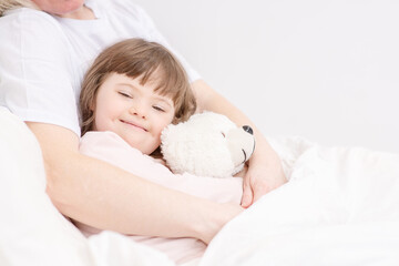 Mom hugging a sleeping girl with down syndrome holding a teddy bear at home in the bedroom on the bed. Ordinary childcare in a family for children with disabilities