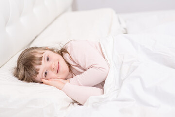 Fototapeta na wymiar A girl with down syndrome lying on the bed under the covers and getting ready for bed. Usually childhood in a family for children with disabilities
