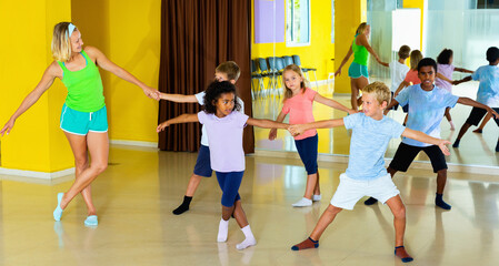 Group of cheerful smiling children practicing vigorous jive movements in dance class with female coach