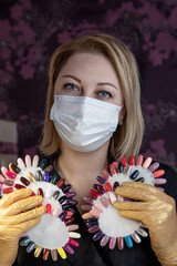 Portrait of a manicurist at the workplace wearing a mask and protective gloves with palettes of multi-colored varnish. Manicurist in a black jacket, protective mask and gloves at the workplace. 