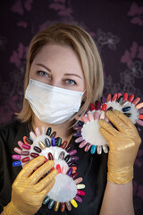 Portrait of a manicurist at the workplace wearing a mask and protective gloves with palettes of multi-colored varnish. Manicurist in a black jacket, protective mask and gloves at the workplace. 