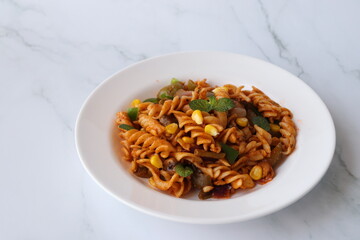 Whole wheat fusilli Pasta Arrabbiata with vegetables. Vegetable Pasta with zucchini, mushrooms, corn, and peppers on a white plate. on a white marble table with cutlery. copy space.