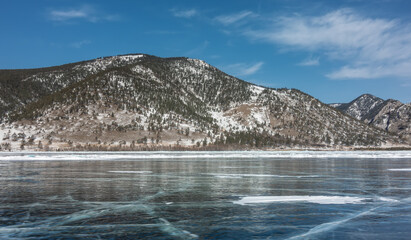 On the smooth shiny surface of the ice of the frozen lake, cracks and areas of snow are visible. On the shore, against the background of the blue sky, there are wooded mountains. Baikal