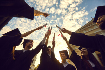 Bottom view graduates lift their diplomas and stack them together against a blue sky with clouds....