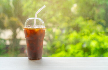 Iced Americano coffee in cleared plastic glass with cover and white straw, coffee glass is put on desk, blurred background of tree, warm light, blank space on front of glass for branding and copy.