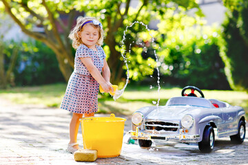 Cute gorgeous toddler girl washing big old toy car in summer garden, outdoors. Happy healthy little...