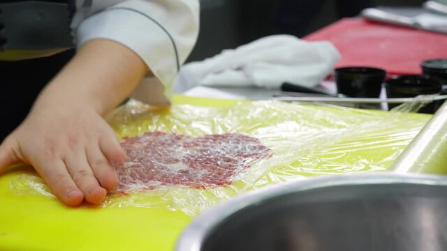 Chef chops raw meat throw food wrap film at commercial kitchen, close-up