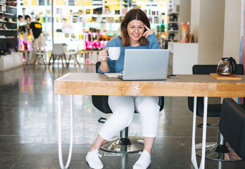Beautiful stylish smiling brunette young woman plus size body positive using laptop at beauty salon...