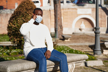 Obraz premium Young African American man sitting on a park bench talking on the phone. Latino man wearing anti-contagion mask. Concept of new normality. 