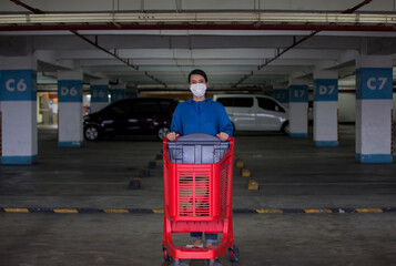 A young woman wears medical face mask against corona virus covid-19 while pushing shopping cart outside of a grocery store in empty parking garage. Food supplies shortage. Panic buying and hoarding.