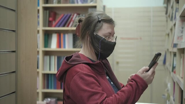 Serious caucasian woman in glasses and a mask against the virus takes photos of bookshelves in a bookstore. Concept of buy literature and gaining knowledge during epidemics and pandemics of virus