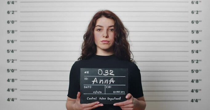 Mugshot of millennial arrested woman with curly hair being photographed in police station. Young female criminal raising head and looking to camera while standing front of metric lineup wall.
