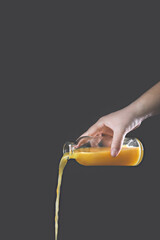 A woman's hand pours orange juice from a glass bottle on a gray background.