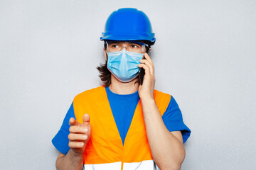 Studio portrait of young worker engineer talking by smartphone, wearing medical face mask against coronavirus or covid-19, and safety construction equipment on grey background.