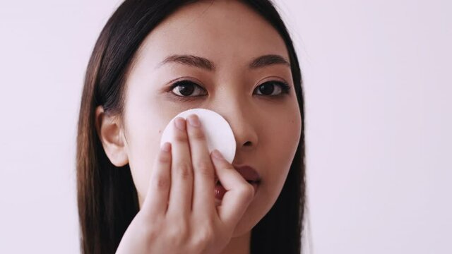 Korean facial cleansing. Skincare hygiene. Beauty routine. Portrait of Asian woman removing nude makeup from smooth skin using cotton pad micellar water isolated on light copy space background.