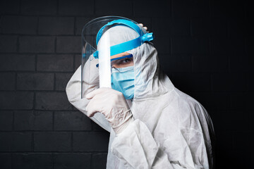 Side portrait of young doctor wearing PPE suit against coronavirus and covid-19, on the background of black brick wall.