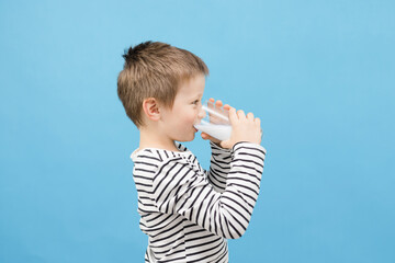 Cute caucasian boy drinks milk or kefir from a glass glass on a blue background. The benefits of...