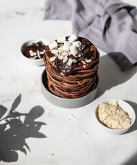 Stack of homemade chocolate pancakes with melted chocolate, ground almond and marshmallow, light background. Direct sunlight and hard shadows.