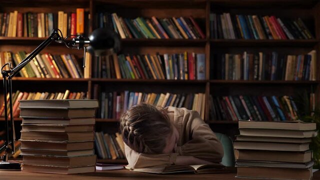 Tired schoolgirl sleeps putting head on copybook among book stacks on wooden table against bookcase under electric light in late evening at home