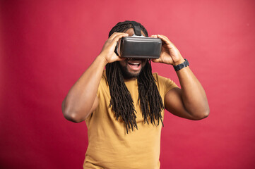 Black happy man with long dreadlocks dressed in casual clothes, standing isolated over red background with virtual reality headset, glasses in an empty room, traveling online, trying new experience