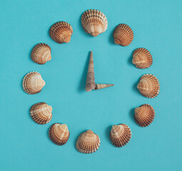 A clock made of seashells lie down on pastel blue background. Minimalistic summer composition