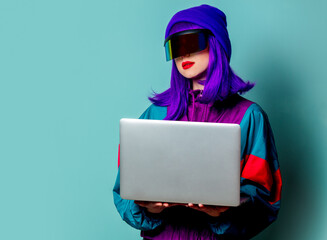 Stylish girl in cyber punk glasses and 80s tracksuit hold laptop computer on blue background