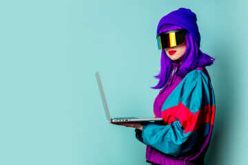 Stylish girl in cyber punk glasses and 80s tracksuit hold laptop computer on blue background