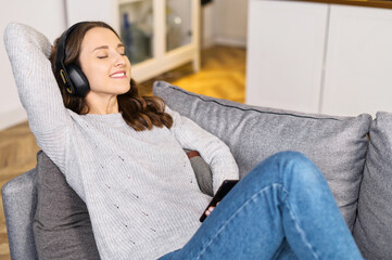 Cheerful serene young woman wearing headphones enjoys favorite tracks, relaxed lying down with eyes closed on the sofa, resting, listening calm music and daydreaming, spends lazy weekend