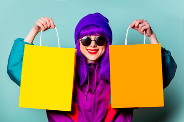 Stylish shopaholic girl in 80s tracksuit and sunglasses hold shopping bags on blue background