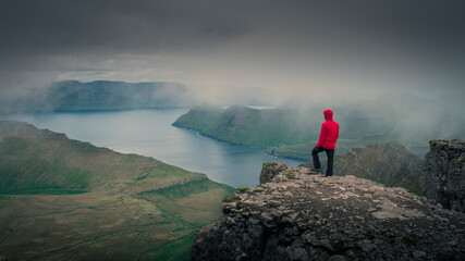 Man in red jacket overlooking green mountain panorama with view to fjord from highest mountain summit on Faroe Islands, Slættaratindur on island Eysturoy.