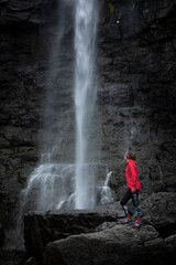 Woman stands on rock with magenta jacket, looks at Fossa waterfall on Streymoy Island, Faroe Islands.