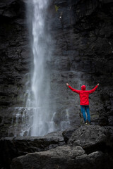 Man stands on rock with red jacket, looks at Fossa waterfall on Streymoy Island, Faroe Islands.