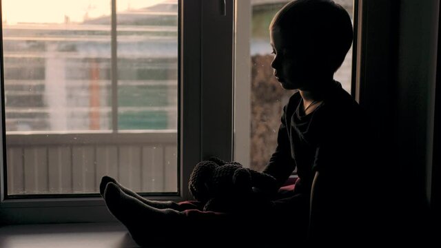 Quarantine, threat of coronavirus. Sad child and his teddy bear. Side view small caucasian boy sitting by window and looking outside. Child staying at home and feeling bored during pandemic.
