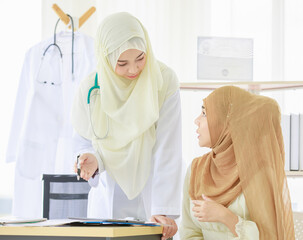 Fototapeta na wymiar Portrait shot of adult Muslim doctor wearing hijab sitting at the table, talking to a patient while using a digital tablet. Concept of modern healthcare medicine in a hospital. Book in the background