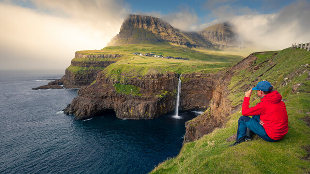 Man with red jacket sits at cliff in front of M&uacute;lafossur waterfall with G&aacute;sadalur village on Vagar island, Faroe Islands.