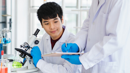 Asian adult man working with teammate standing do writing hold note book both are scientist and wearing uniform and eyeglass. Microscope, test tube with liquid put on table in laboratory