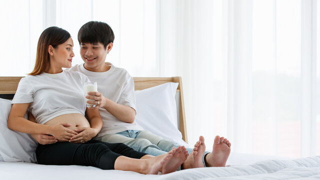 happy and healthy family concept. Picture of a young couple sitting on a bed together. A young husband smiling and holding a glass of milk and giving it to a pregnant wife with love