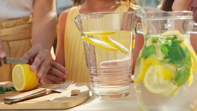 Slow-motion midsection shot of unrecognizable woman and two girls making lemonade outdoors in summer, cutting fresh lemons and putting them in glass decanter