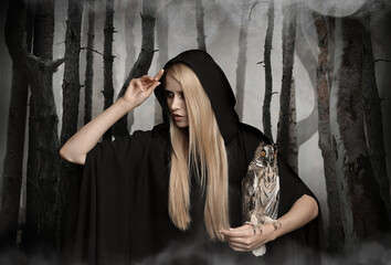 Witch wearing black mantle with owl in foggy forest. Fantasy world